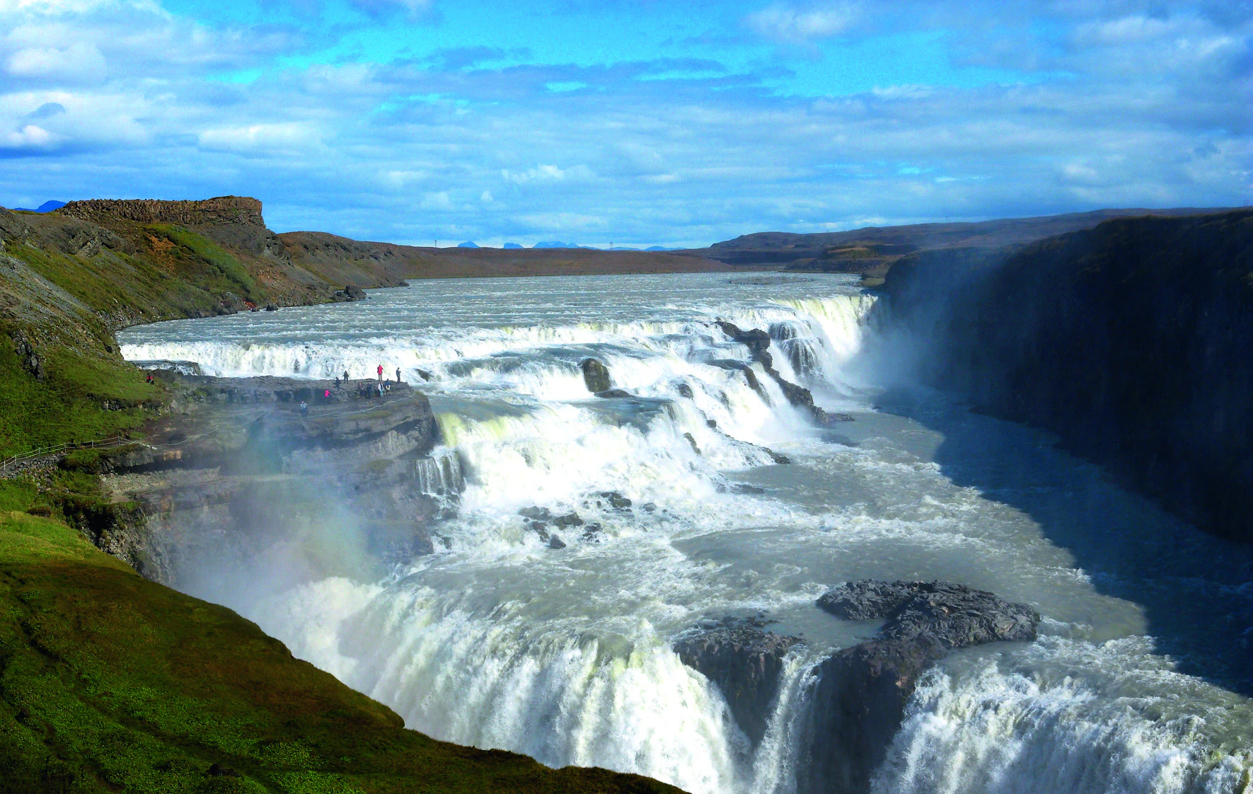 Wide waterfall cascading over multiple levels with rocky terrain, under a partly cloudy sky. Small figures stand on a viewing platform.
