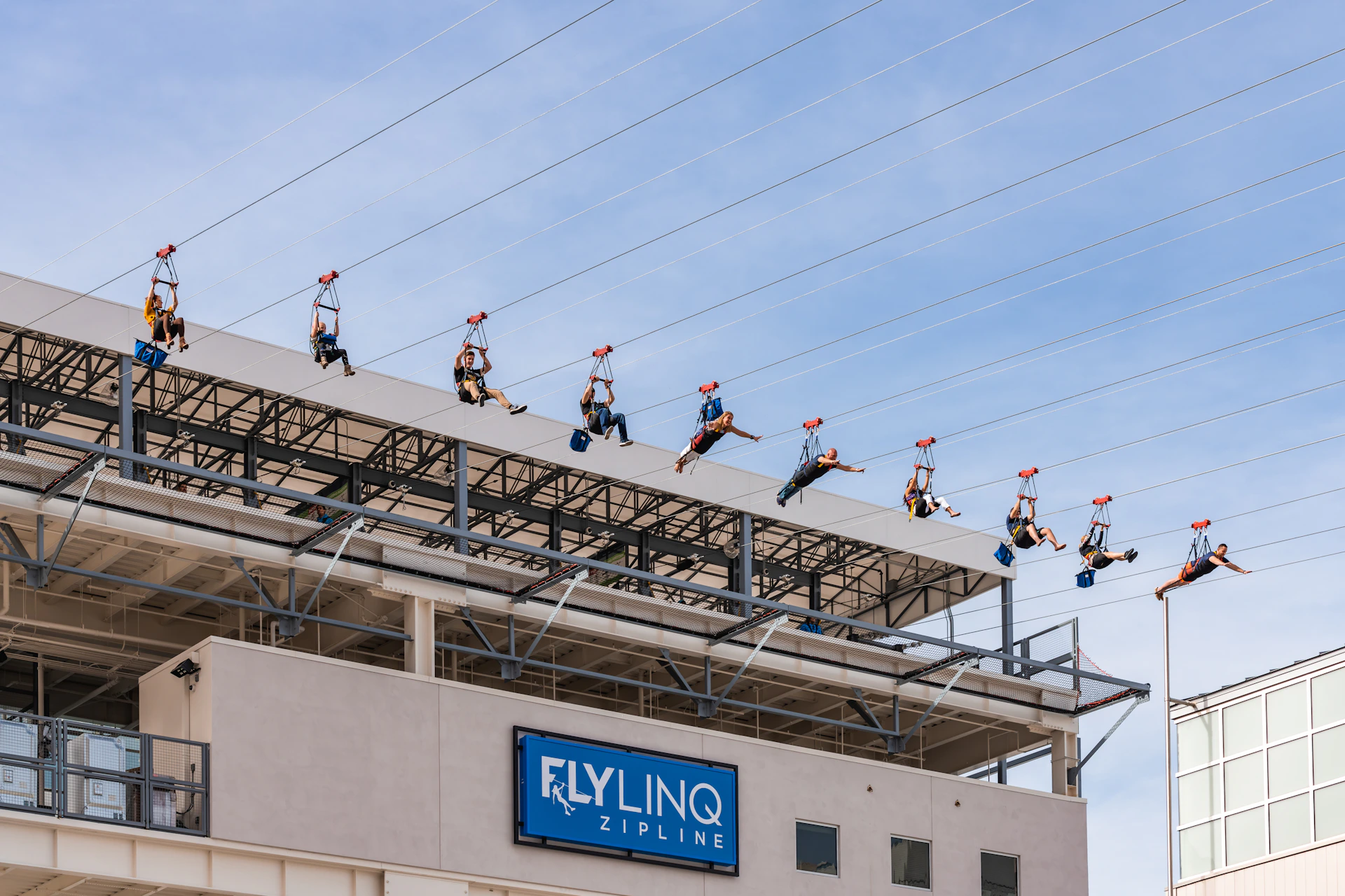People ziplining above a building with a sign that reads "Fly LINQ Zipline" and a clear blue sky in the background.