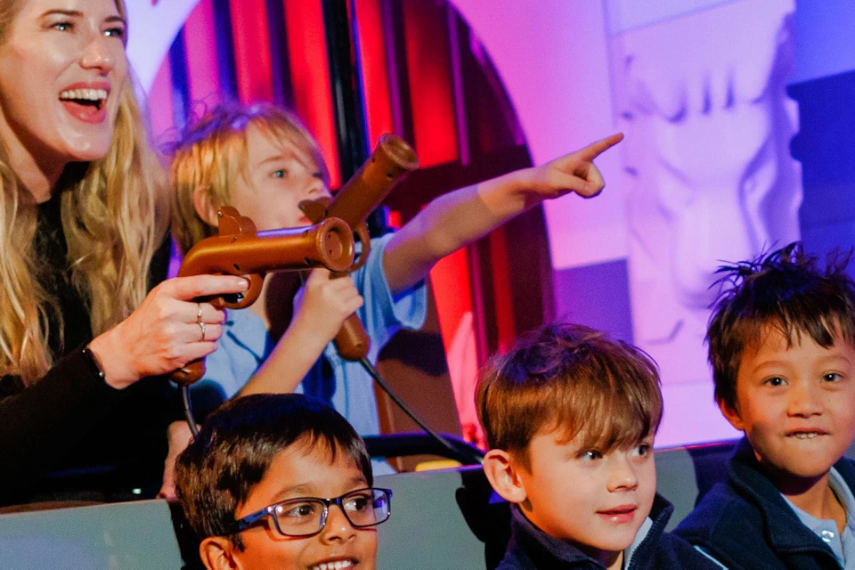A woman and three boys enjoy a ride, holding toy guns and smiling, against a colorful, medieval-themed background.