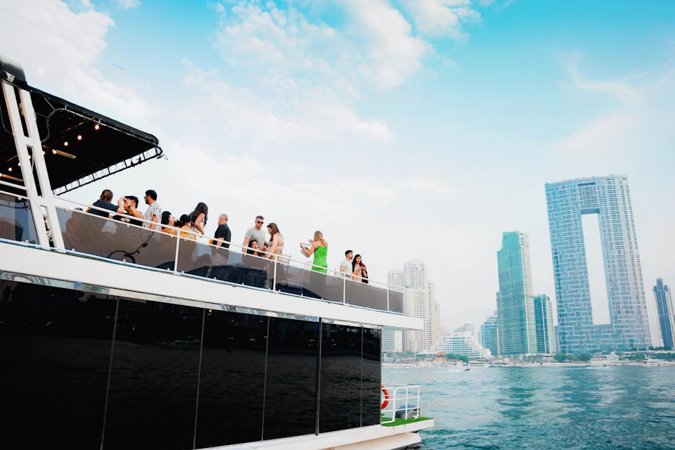 People socializing on the upper deck of a boat with city skyscrapers and a blue sky in the background.
