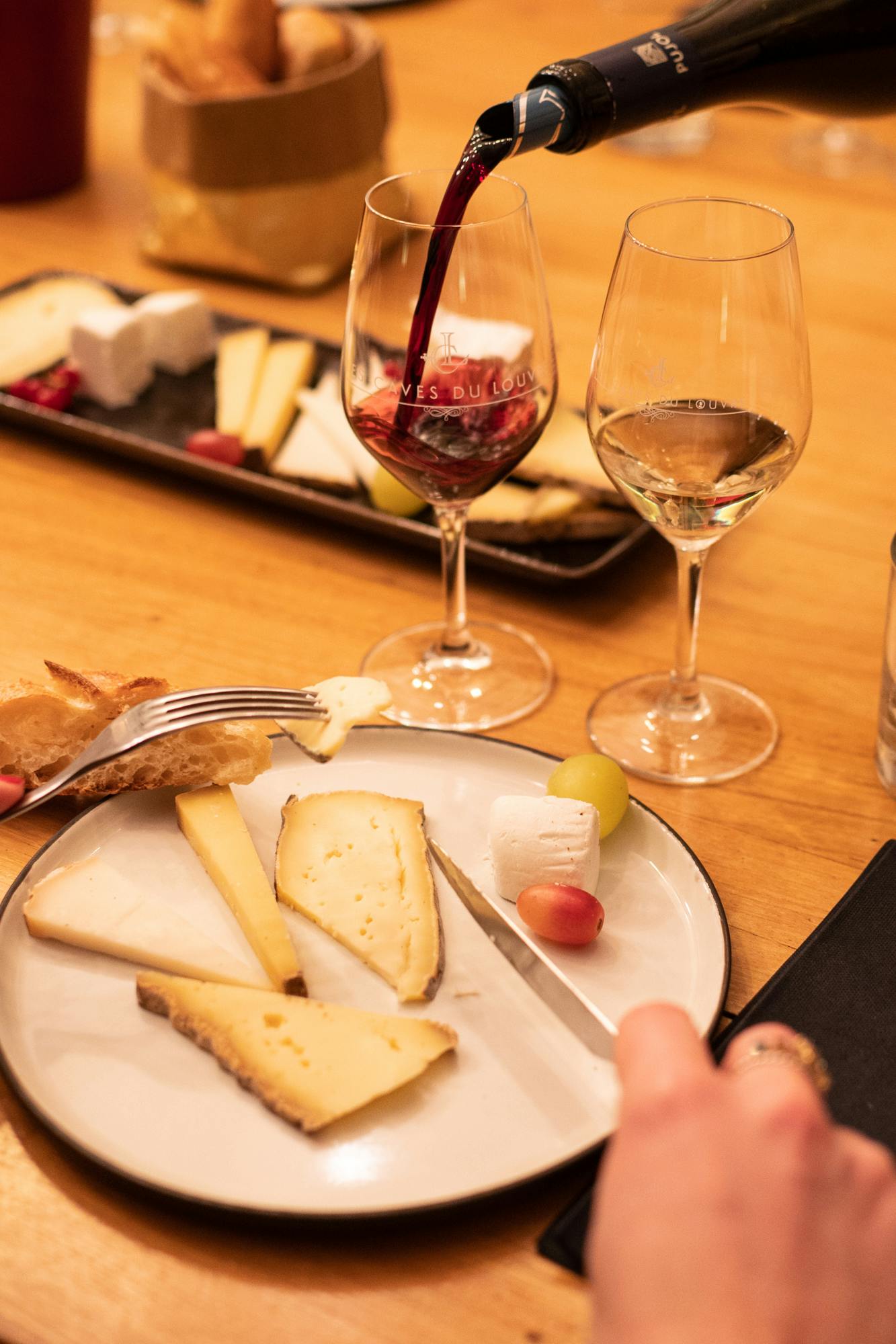 Тарелка с различными сырами, виноградом и хлебом на столе в сопровождении двух винных бокалов, один из которых наполнен красным вином.