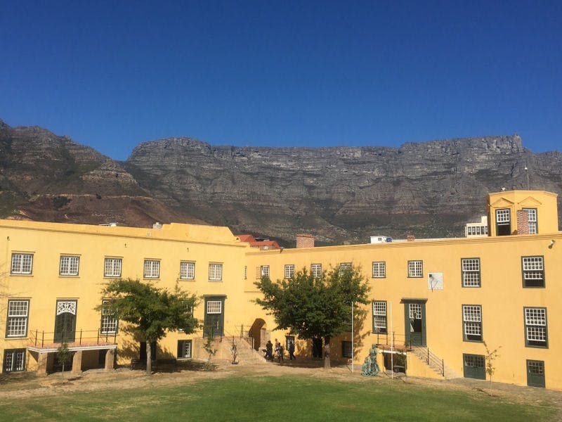 Un edificio giallo con porte e finestre verdi, alberi di fronte e una catena montuosa sullo sfondo sotto un cielo azzurro.
