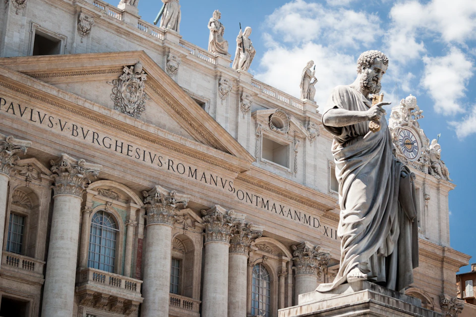Statue holding a key in front of a grandiose building with ornate columns and sculptures on the rooftop.