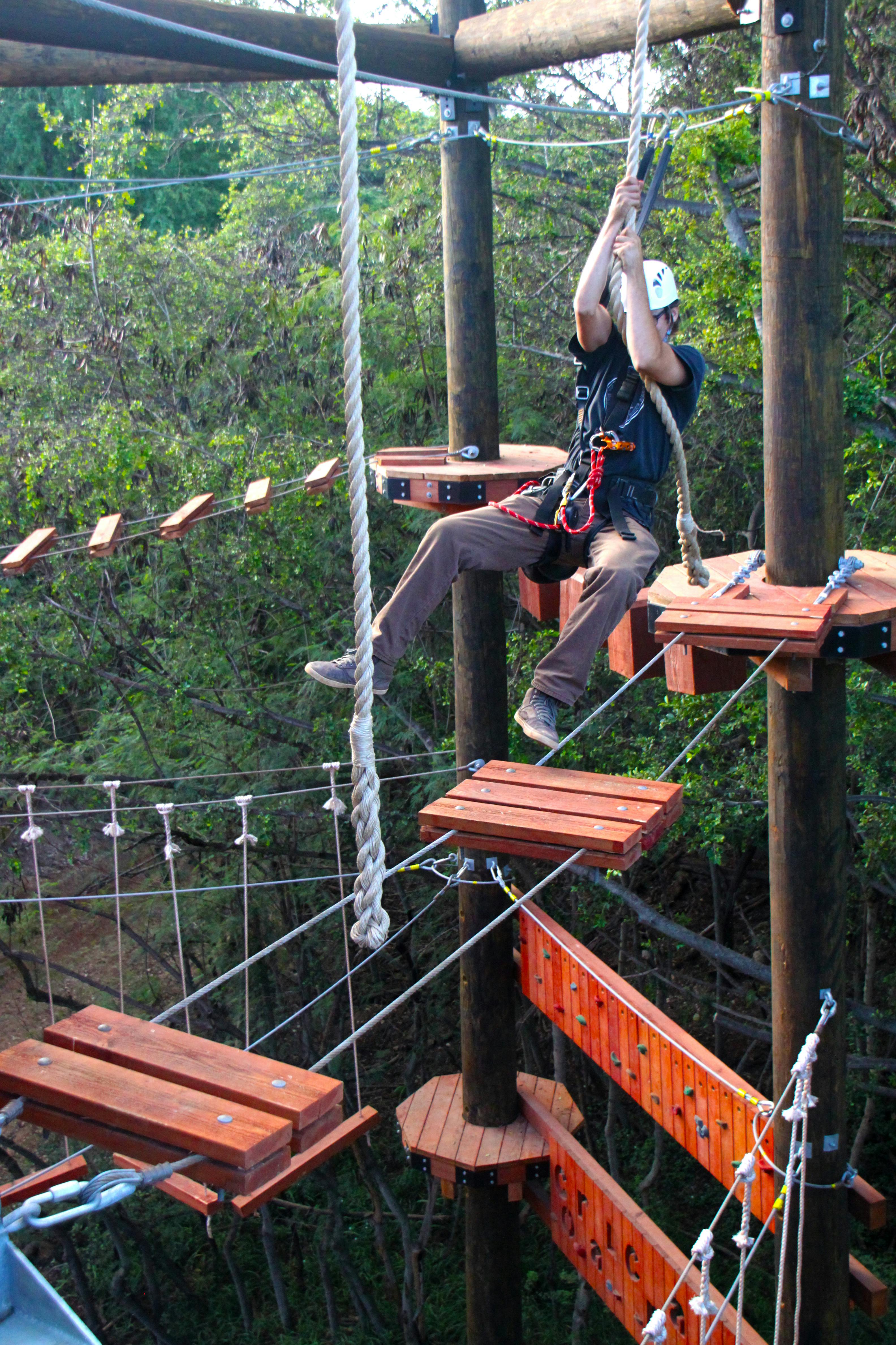 한 사람이 나무로 둘러싸인 안전 장비와 헬멧을 착용하고 나무 플랫폼과 로프가 있는 높은 로프 코스를 항해합니다.