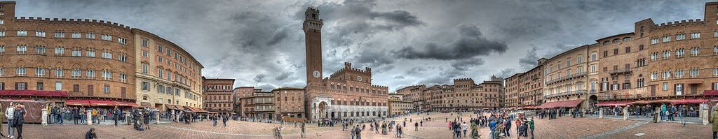 탑이 있는 역사적인 건물로 둘러싸인 넓은 광장의 파노라마 전망, 흐린 하늘 아래, 걷고 모이는 사람들.