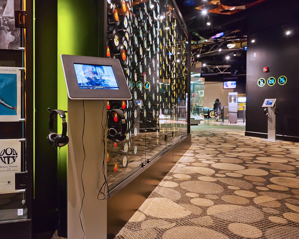 Modern museum exhibit hall with circular-patterned flooring, display of records on the wall, and interactive touchscreen stations.