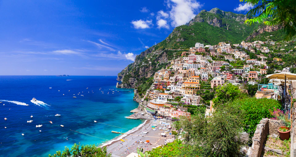A coastal town with colorful buildings on a steep hillside overlooking a blue sea with boats and a beach below.