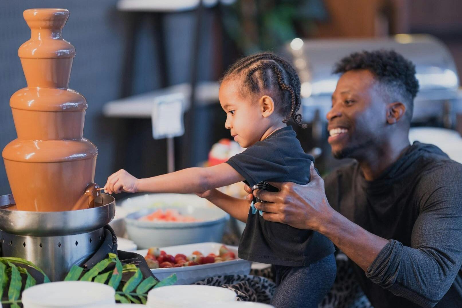 뷔페 테이블에서 음식을 먹을 때 웃는 남자가 어린 아이를 안고 있으며, 과일 쟁반이 배경에 보입니다.