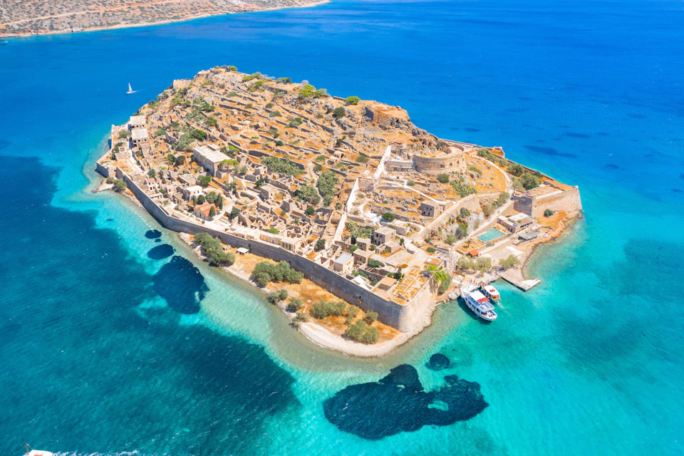 Aerial view of a fortified island with ruins, surrounded by turquoise water. Two boats are docked at the shore.