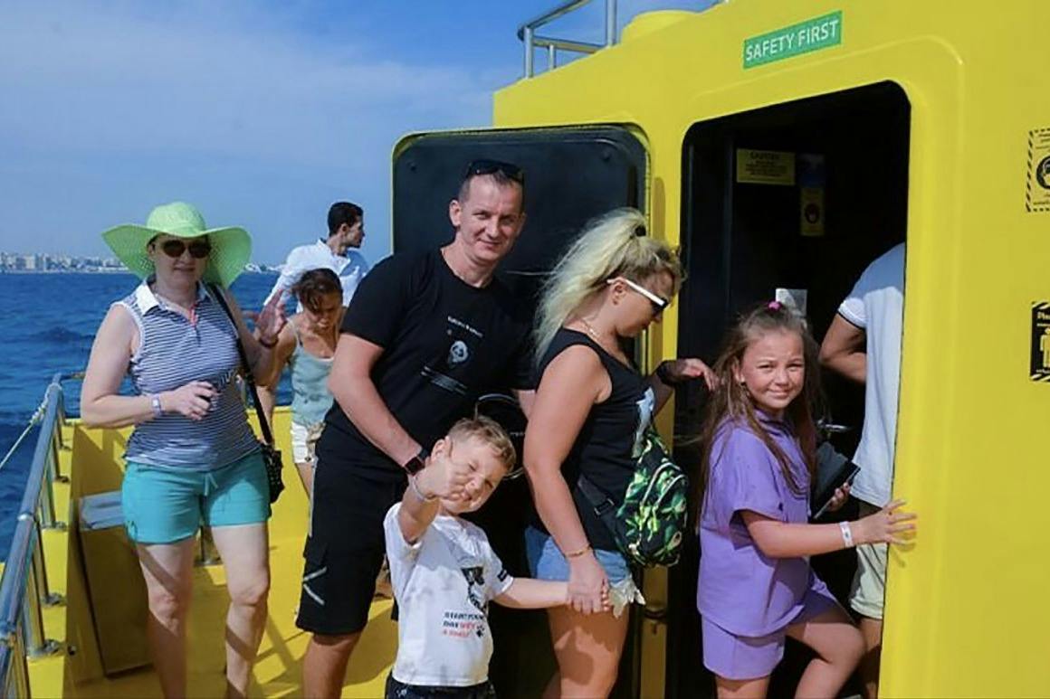 A group of people boarding a yellow vehicle under a sunny sky. A child waves at the camera in the foreground.