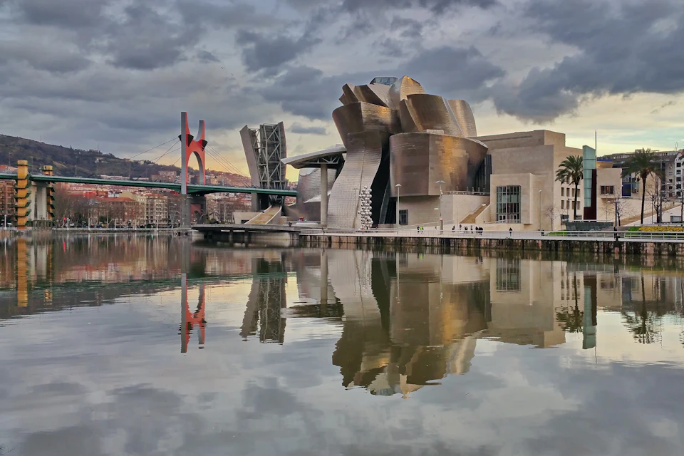 Modern, metallic building reflecting in calm water, with a red bridge and cloudy sky in the background. Sparse pedestrians along the water.