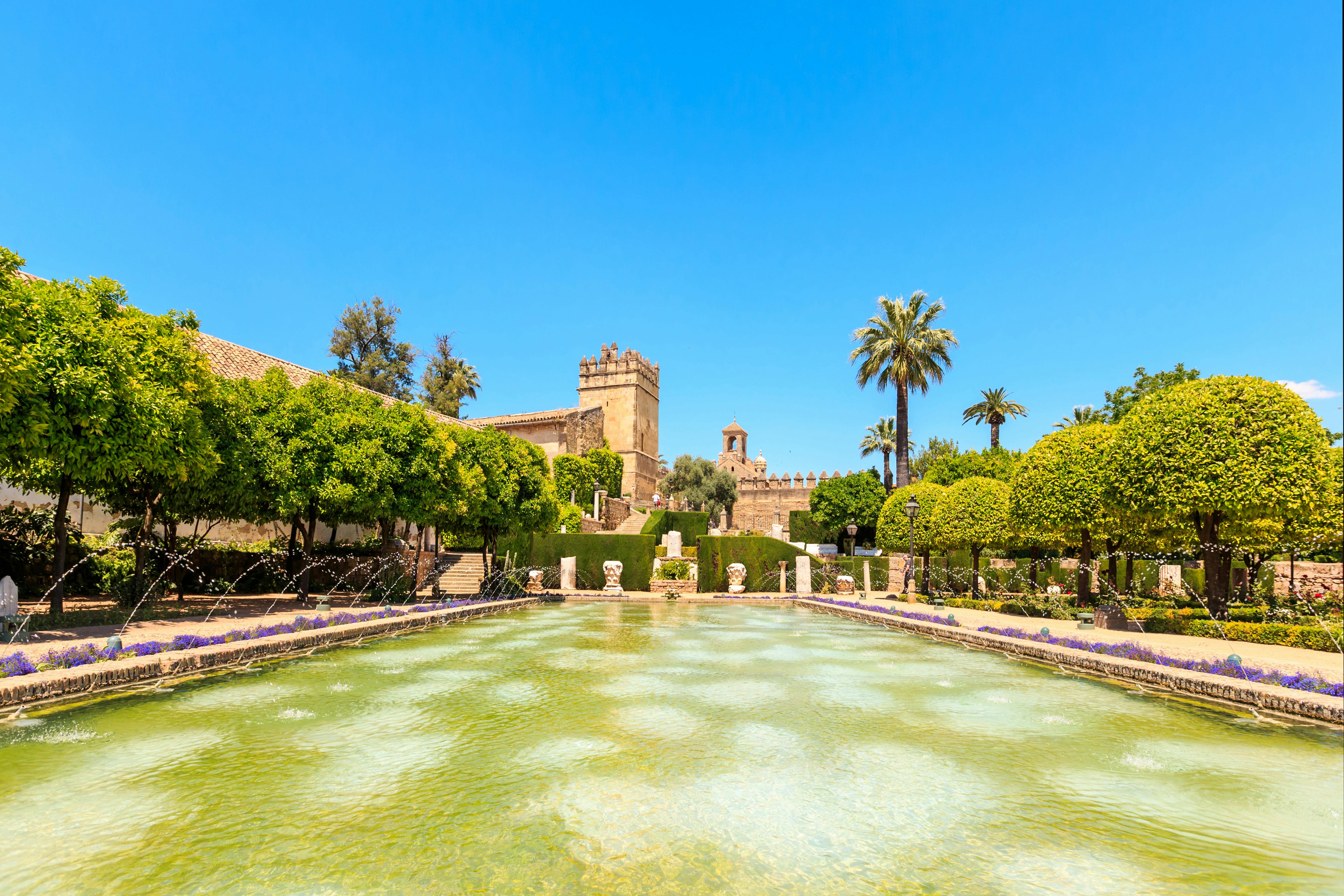 Historic garden with a clear pond, fountain jets, manicured bushes, palm trees, and stone buildings under a bright blue sky.