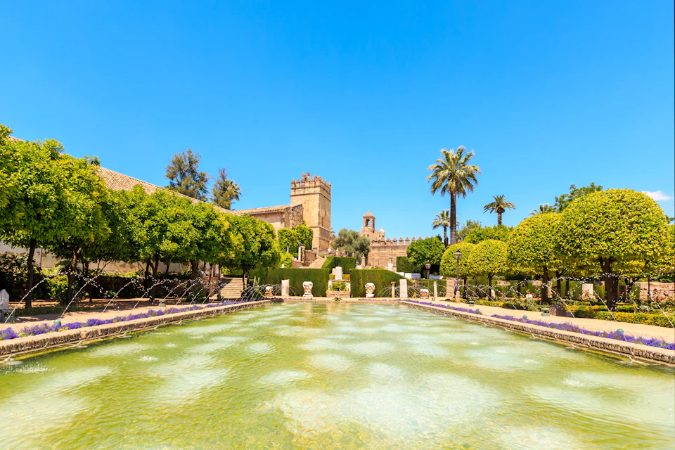 Historic garden with a clear pond, fountain jets, manicured bushes, palm trees, and stone buildings under a bright blue sky.