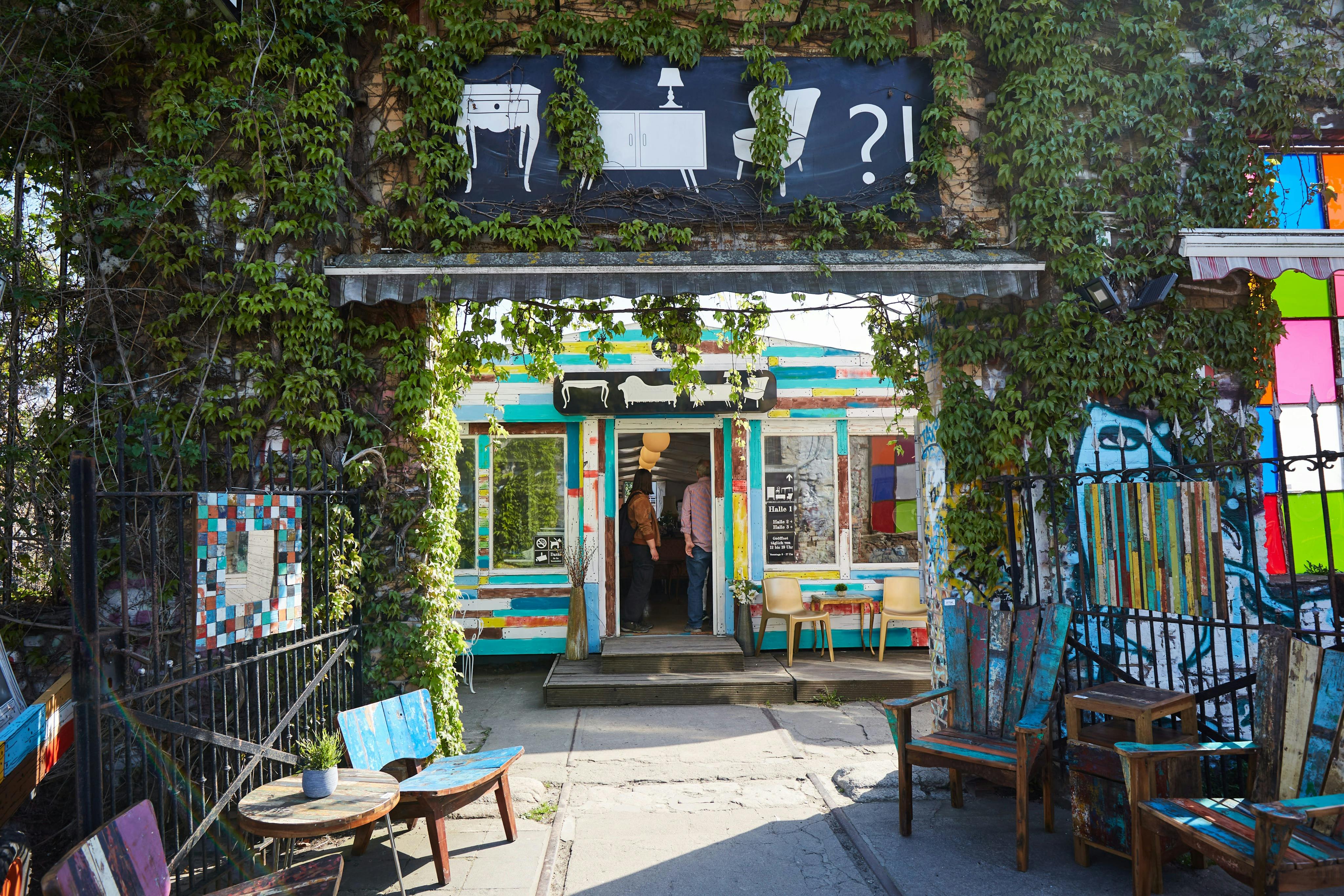 Μια πολύχρωμη βιτρίνα με μια πινακίδα από πάνω, που περιβάλλεται από πράσινα αμπέλια, υπαίθρια καθίσματα και ανοιχτή είσοδο σε εσωτερικό χώρο.