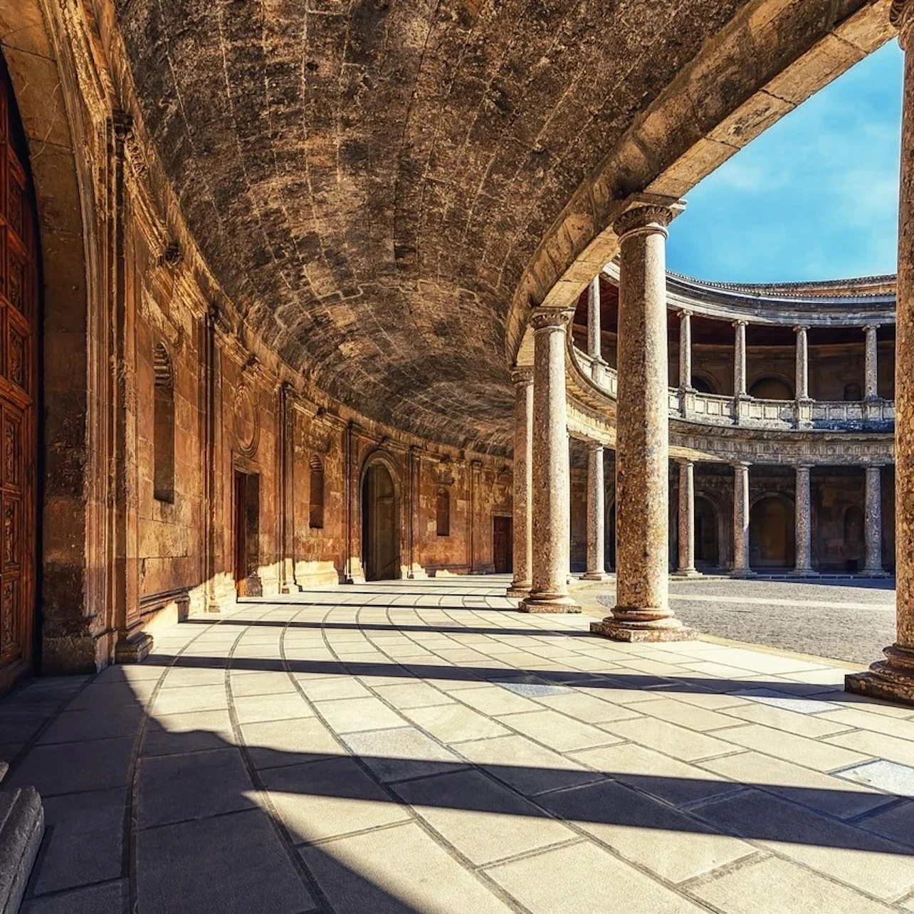 A circular colonnade with tall stone columns and arched architecture, under a blue sky, partially illuminated by sunlight.