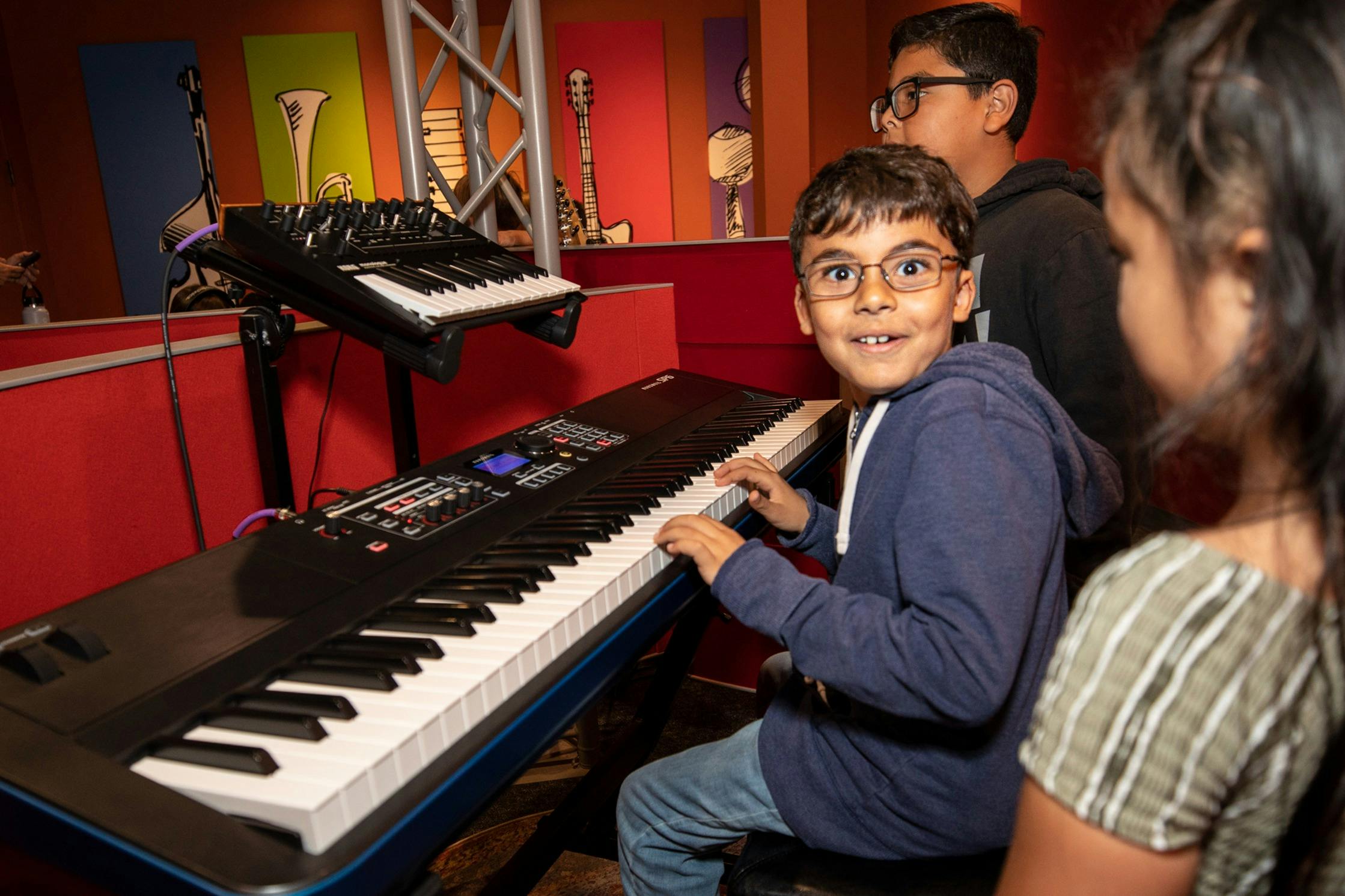 A young boy plays a keyboard with two other children nearby, all smiling, in a room with colorful walls and musical artwork.