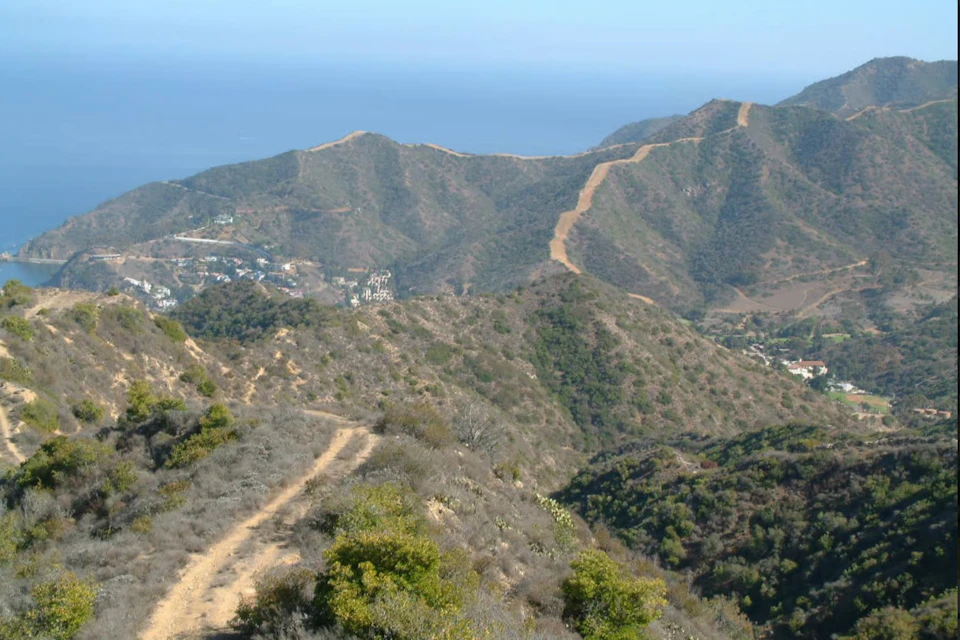 Hiking trail winding through dry, hilly terrain with sparse vegetation, distant mountains, houses, and ocean visible in the background.