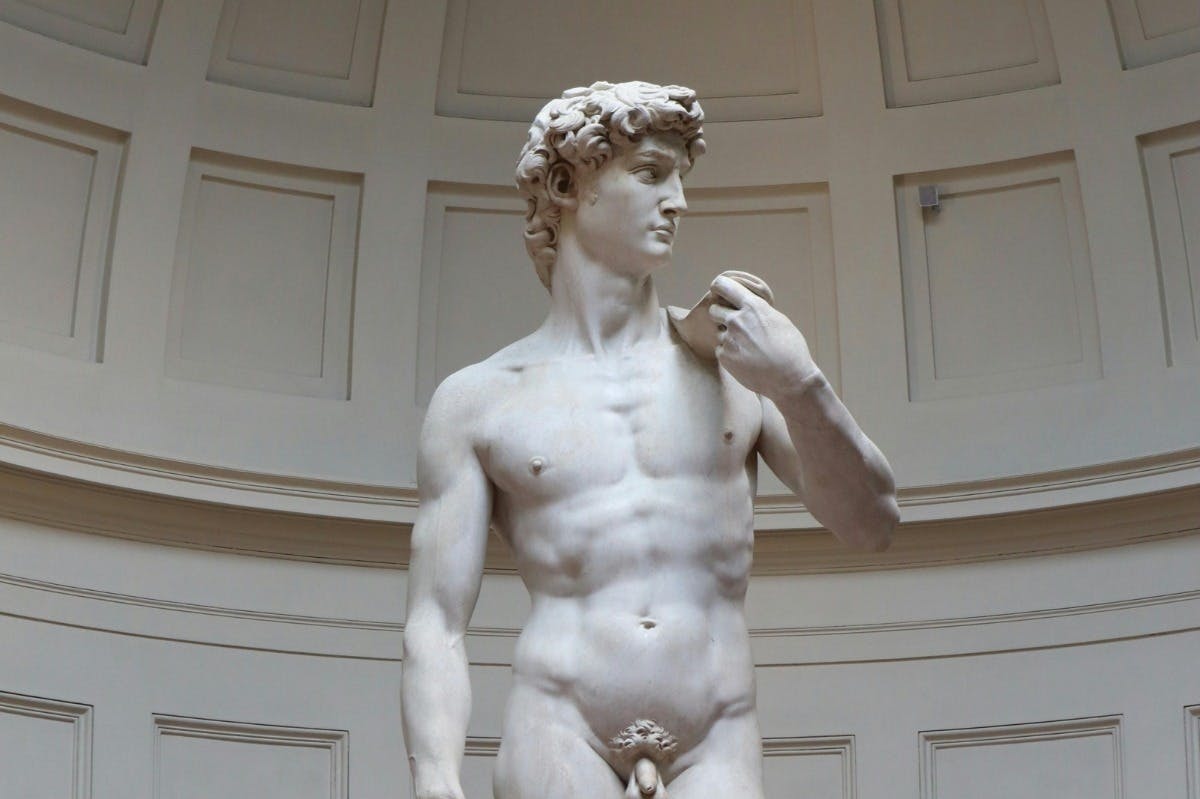 Statue en marbre représentant un homme nu debout, aux cheveux bouclés, tenant un objet sur son épaule à l'intérieur d'un bâtiment orné.
