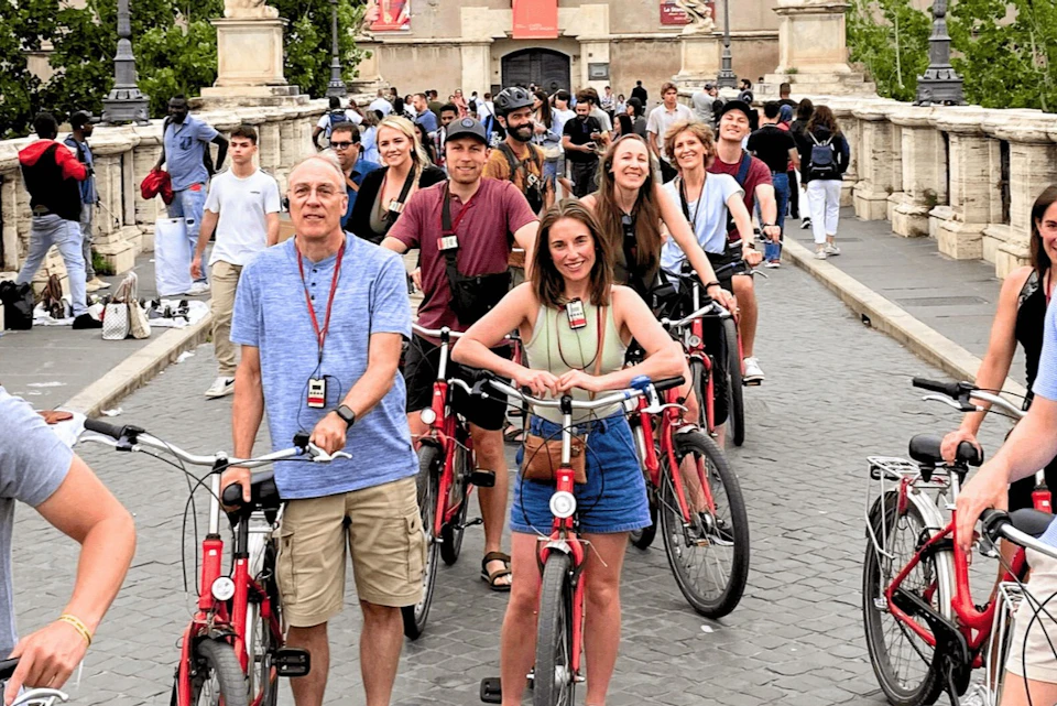 Group of people on red bicycles smiling and posing on a cobblestone path with crowds and historic architecture in the background.