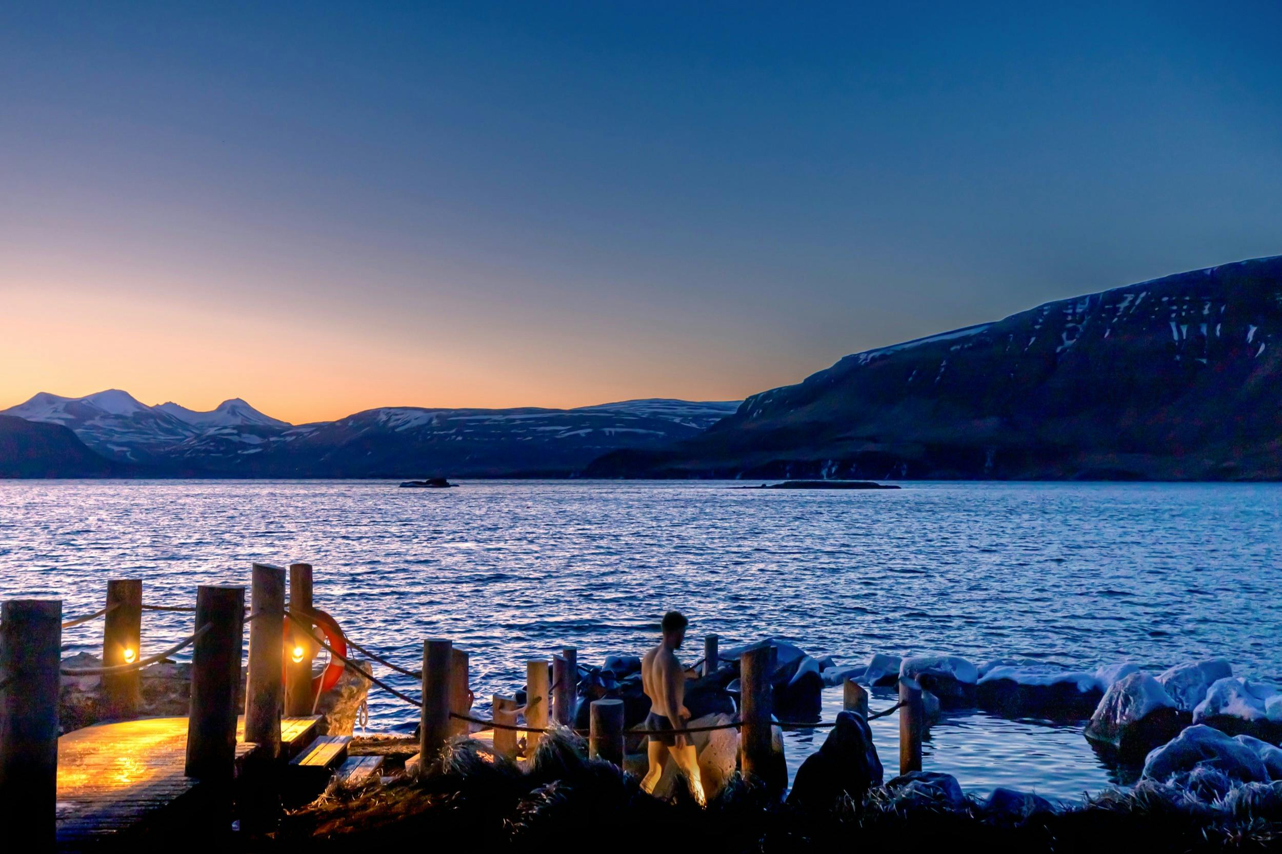 黎明时分，一个人在湖边码头附近散步，背景是山脉的轮廓，渐变的天空从橙色到蓝色不等。