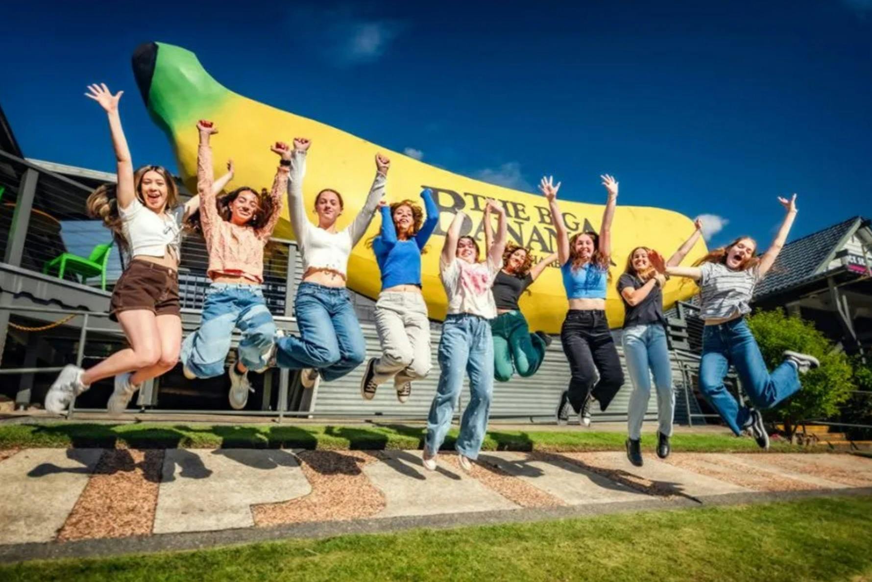En gruppe mennesker hopper foran en stor bananskulptur under en klar blå himmel.