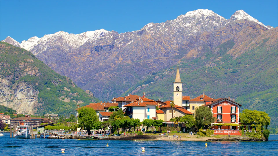 A picturesque lakeside village with red-roofed buildings, a church tower, and snow-capped mountains in the background.