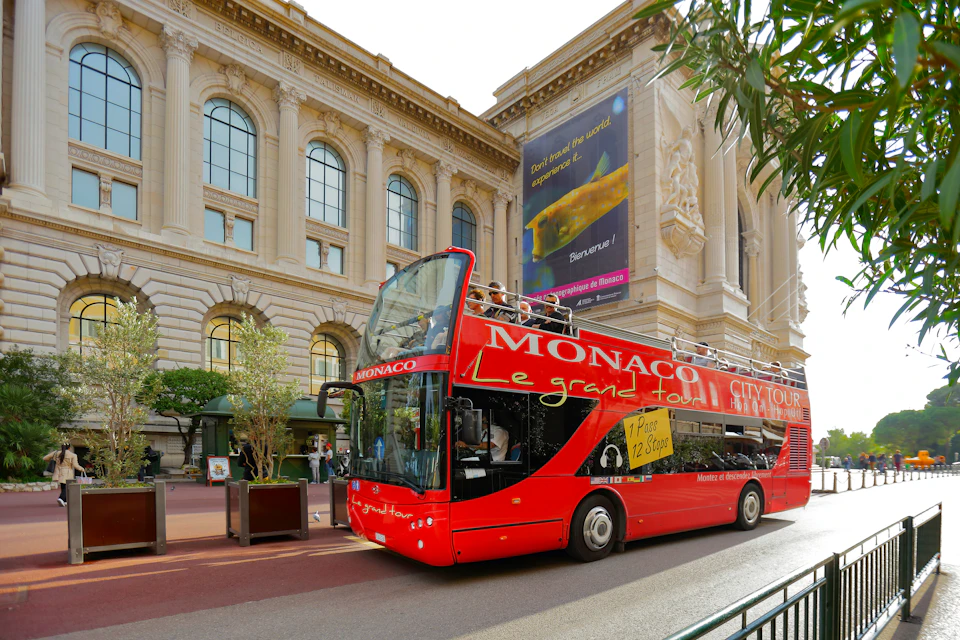 Red double-decker tour bus labeled "Monaco Le Grand Tour" in front of a large classical building with arched windows.