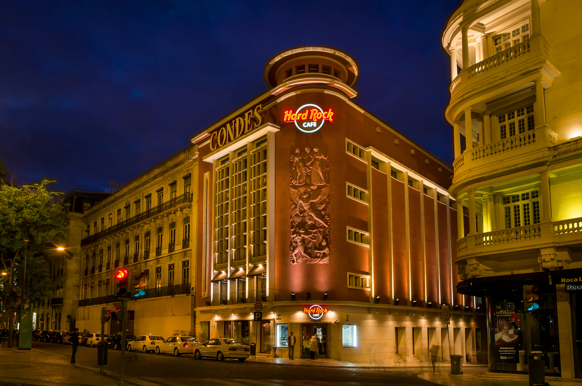 Illuminated Hard Rock Café with "CONDES" sign at night, housed in a historic building with intricate facade details and parked cars outside.