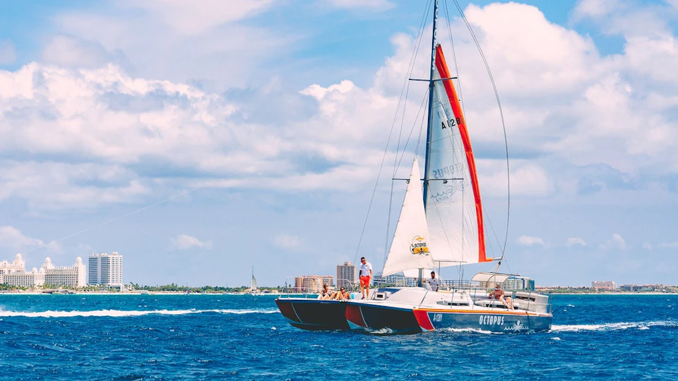 A sailboat with several people aboard is sailing on a blue ocean under a partly cloudy sky, with a city skyline in the background.