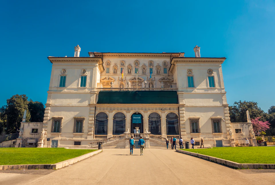 People walk towards a large, ornate building with sculptures and archways under a clear blue sky.