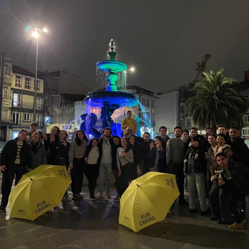 A large group of people pose in front of a lit fountain at night, holding yellow umbrellas with "PUB CRAWL" text.