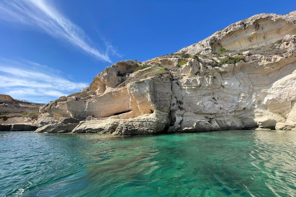 Rocky cliffside with greenish-blue clear water below and a bright blue sky above.