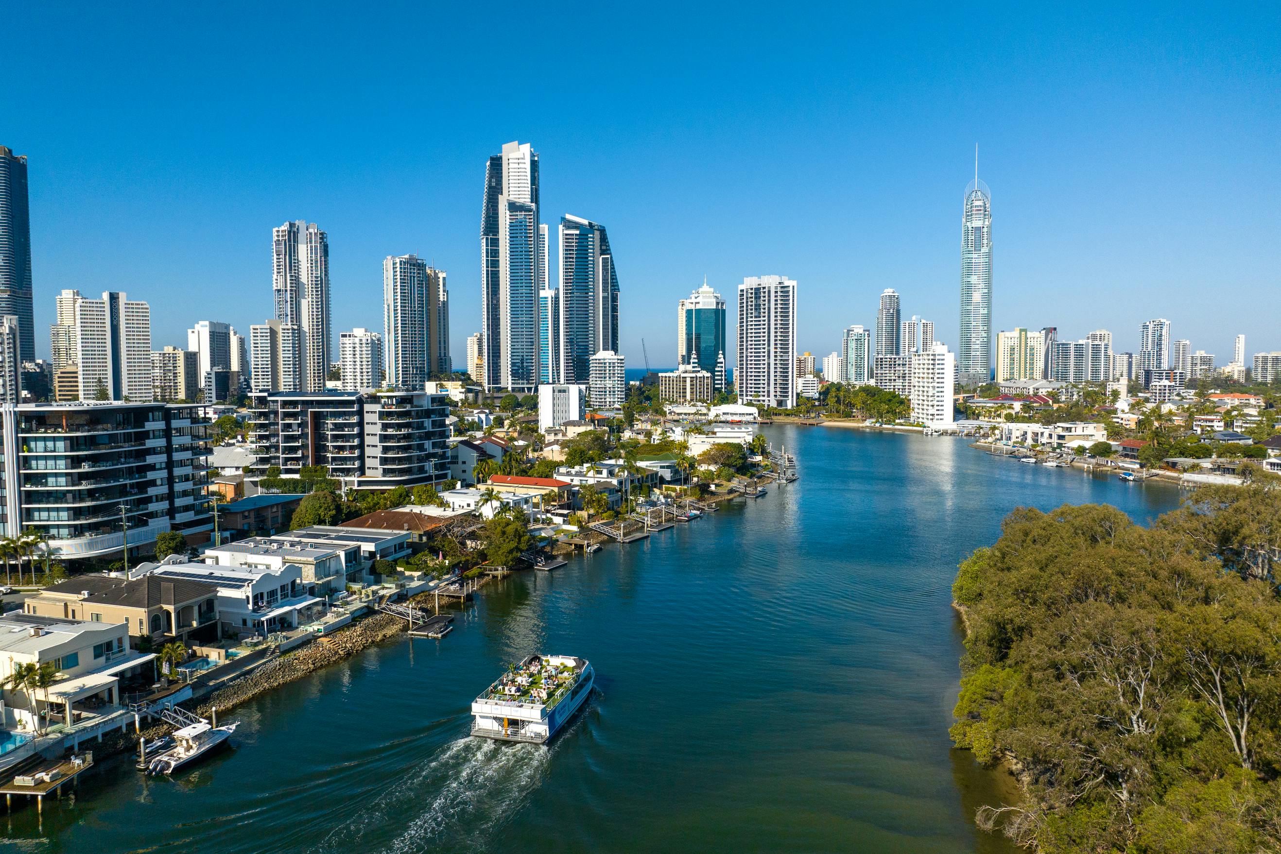 Εναέρια άποψη μιας πόλης με ψηλά κτίρια δίπλα σε ένα ποτάμι, μια βάρκα που ταξιδεύει στο νερό κάτω από έναν καταγάλανο ουρανό.