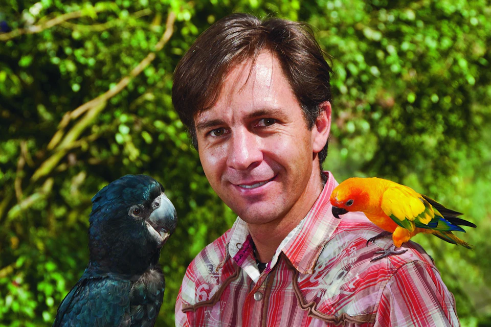 A man standing outdoors with a black parrot on his left and a yellow-orange parrot on his right shoulder.