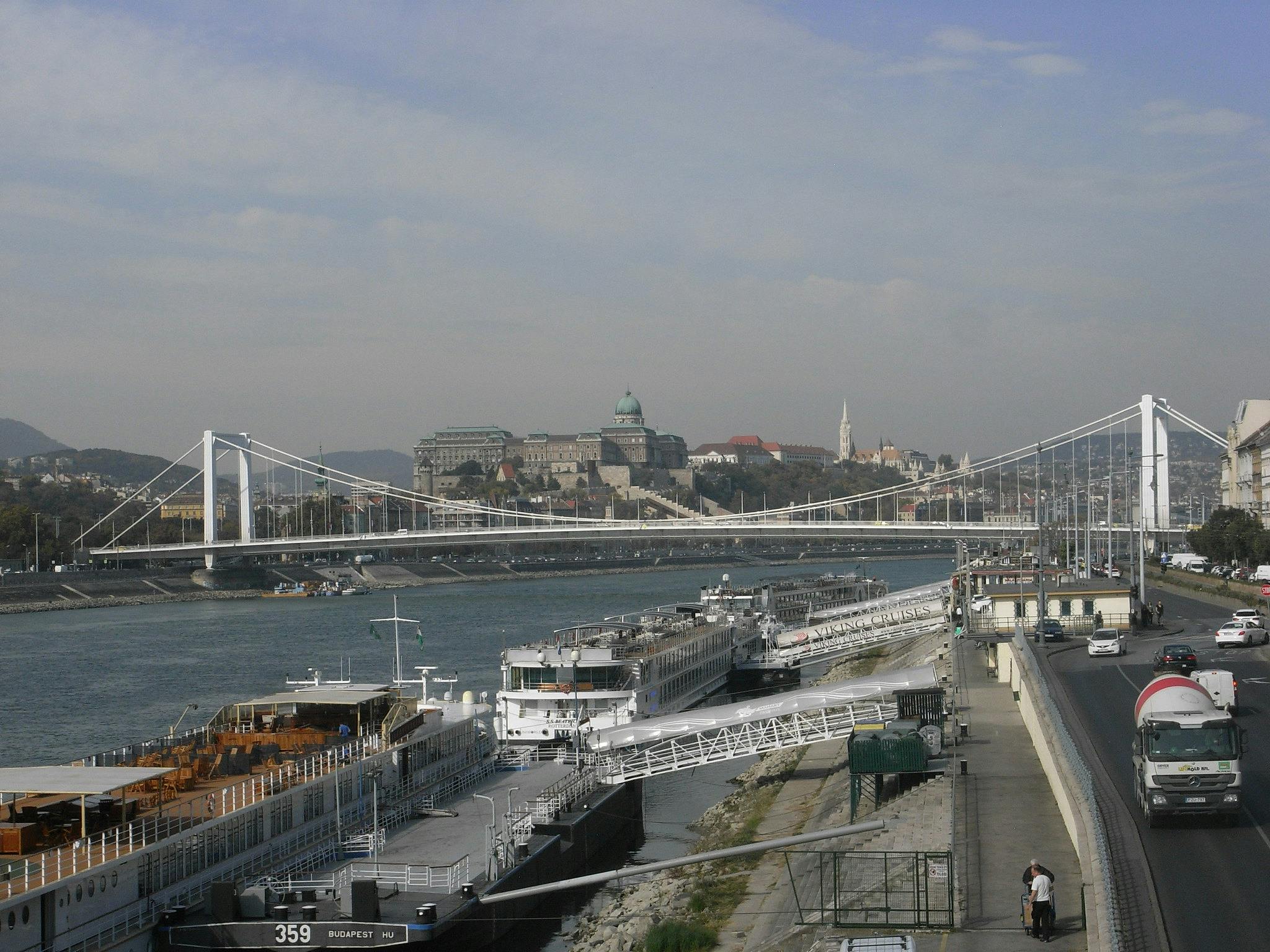Ponte sospeso su un fiume con navi da crociera attraccate. Edifici storici e traffico pedonale sulla strada parallela al fiume.