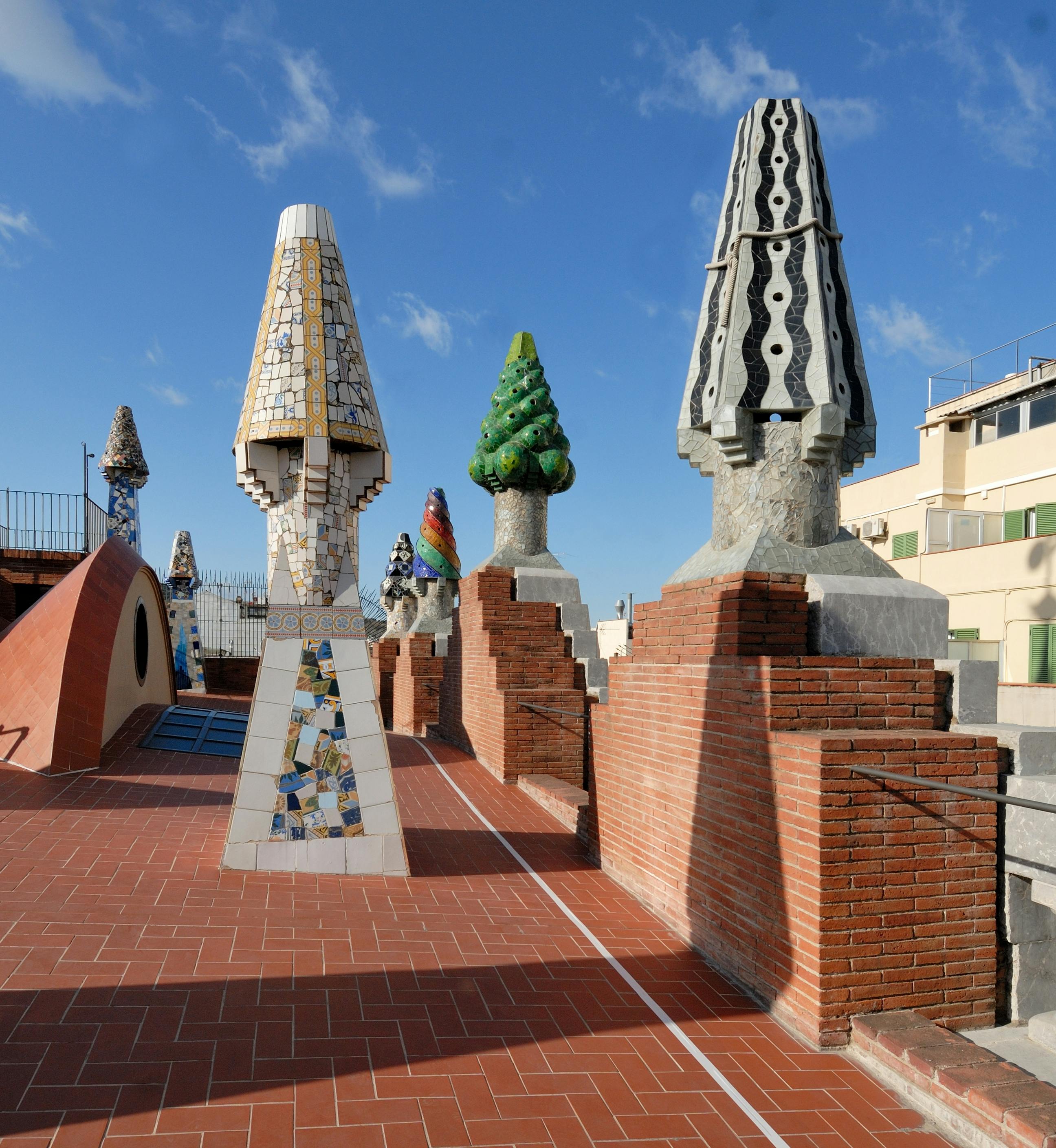 Colorful, abstract chimneys on a rooftop terrace with brick flooring, under a blue sky with a few clouds.