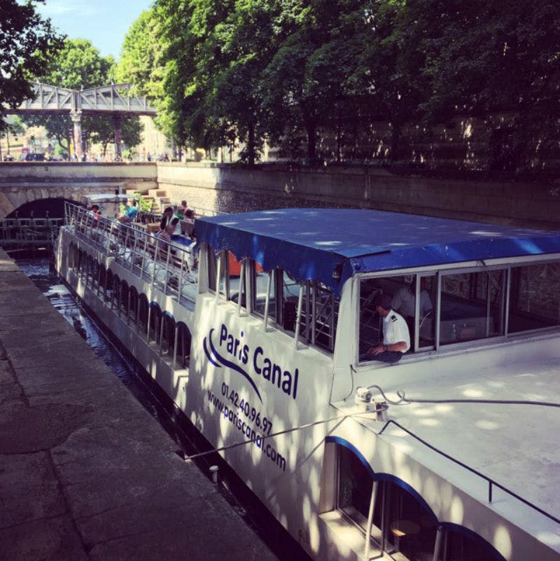 Vyhlídková loď s názvem „Pařížský kanál“ proplouvá kanálem ohraničeným stromy a kamennými zdmi pod mostem.