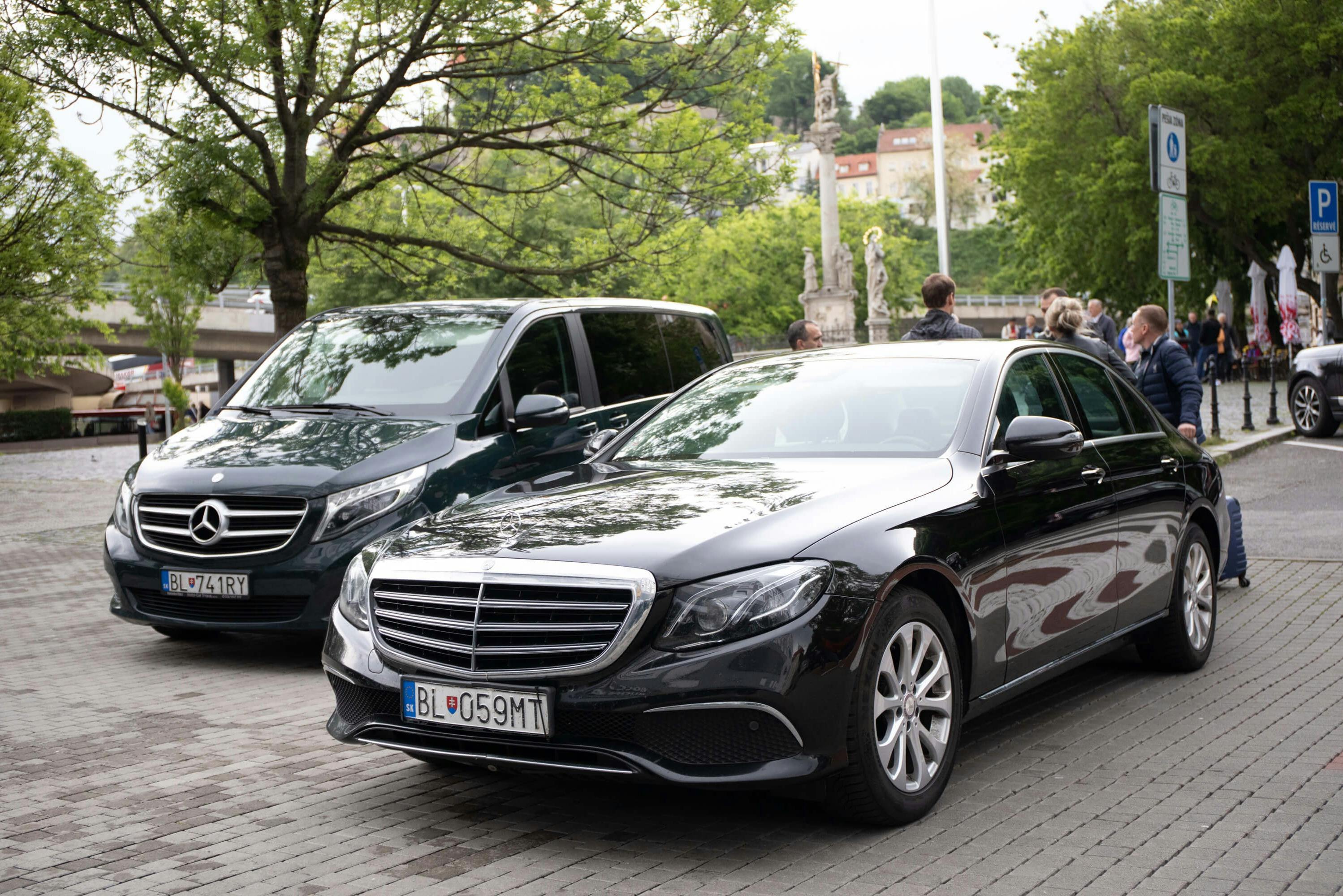 两辆黑色豪华轿车停在鹅卵石街道上，后面有树木和人。