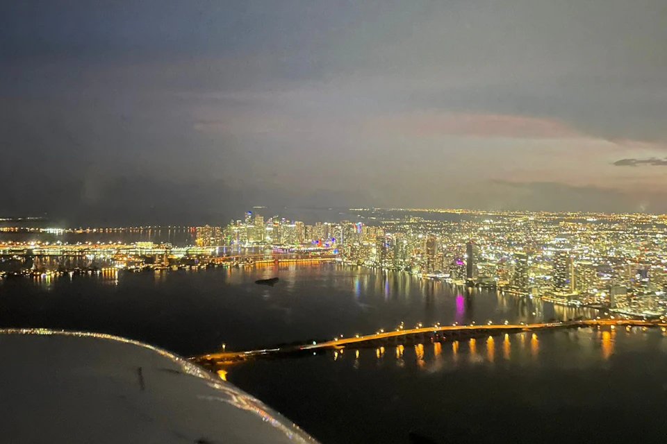 Aerial view of a city skyline at night with illuminated buildings, a bridge over water, and reflections of lights on the surface.
