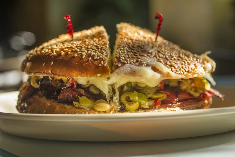 A sandwich with sesame seed buns, melted cheese, sliced olives, and other fillings, secured with red picks on a plate.