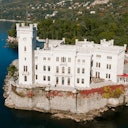 White stone castle situated on the edge of a cliff by a lake, surrounded by dense forest and hills.