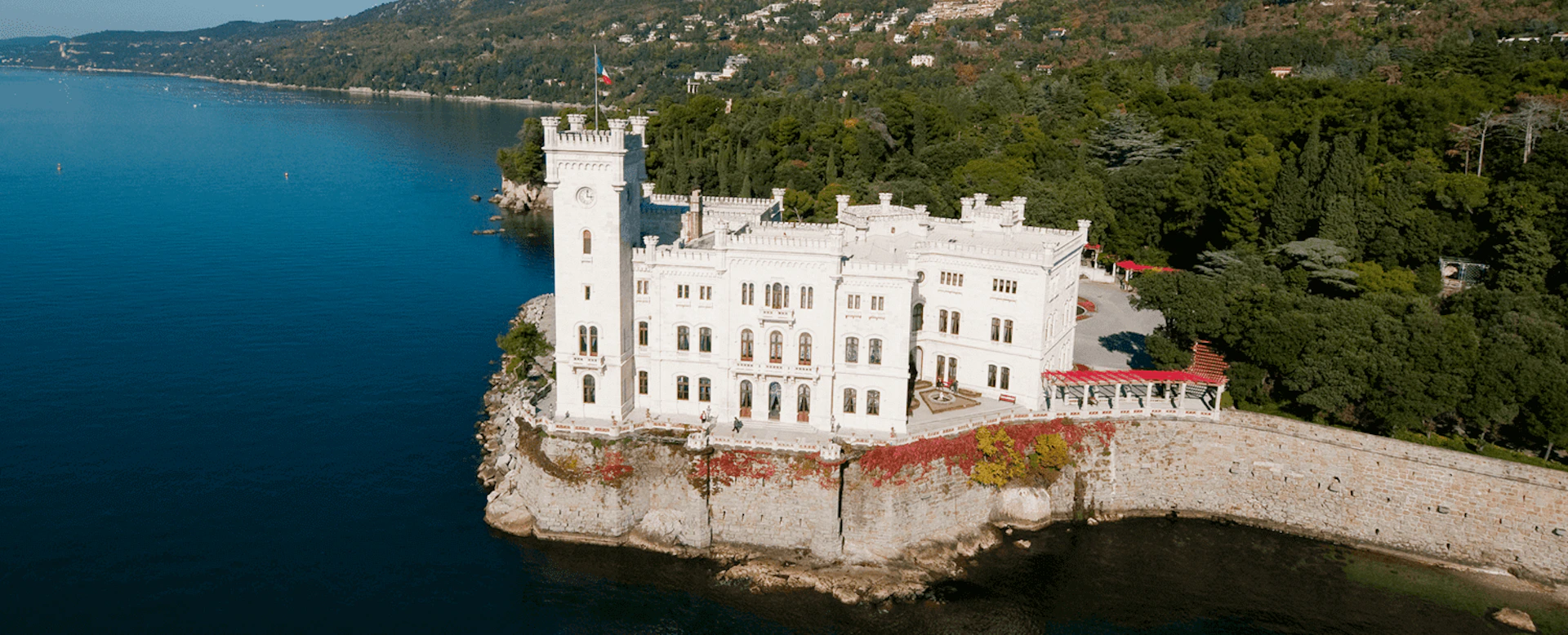 White stone castle situated on the edge of a cliff by a lake, surrounded by dense forest and hills.