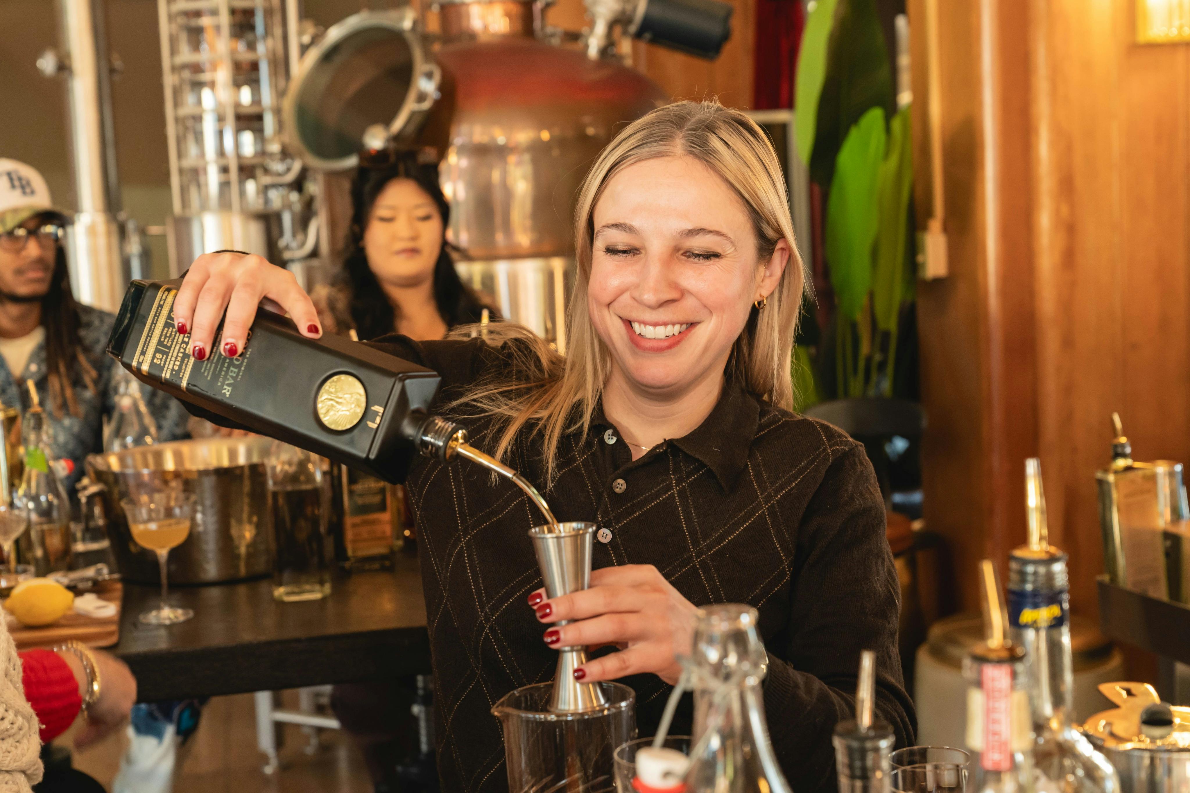 A woman smiles while pouring liquid from a bottle into a jigger, surrounded by others in a distillery setting.