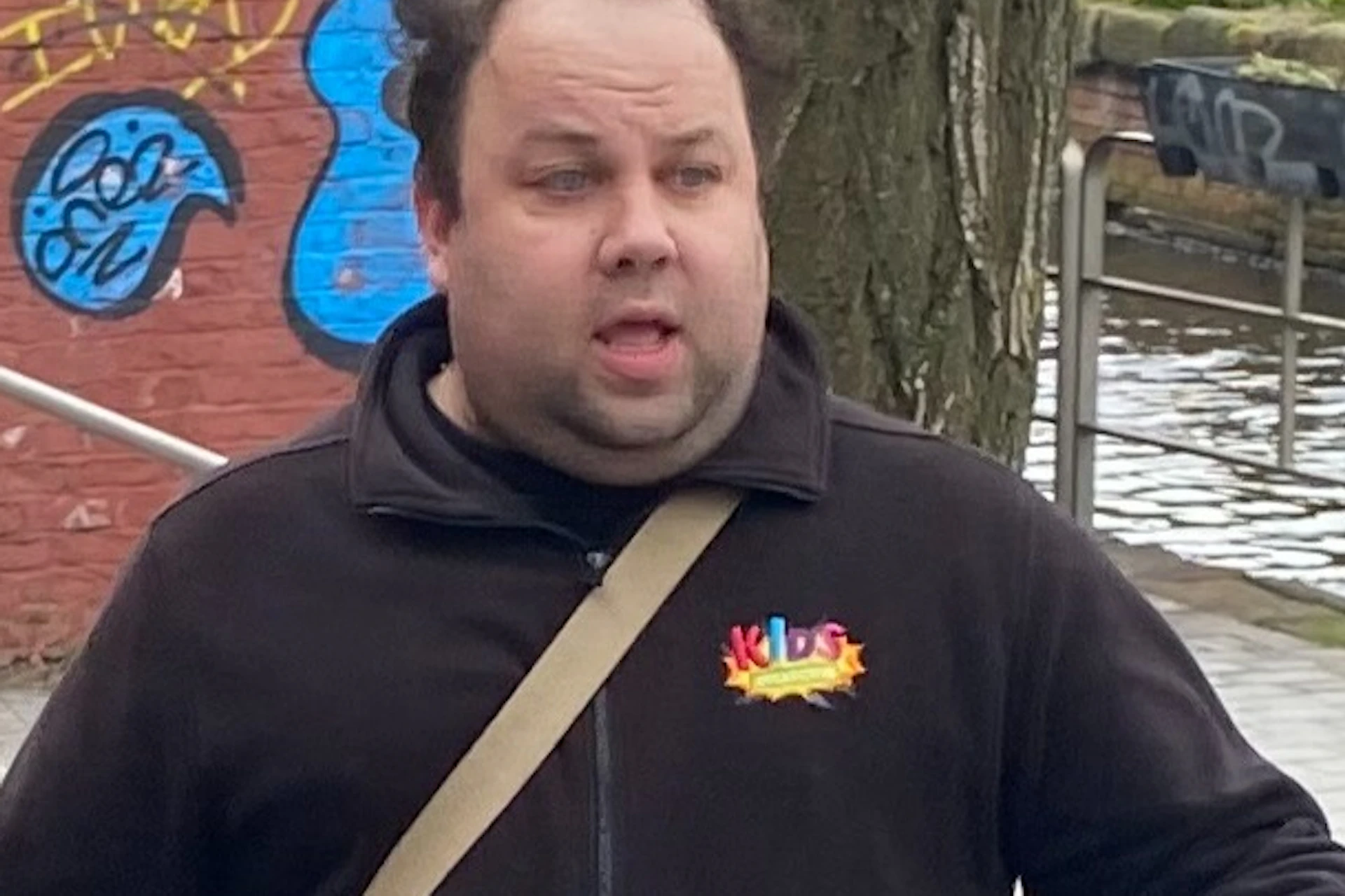 A man speaks to a small group of people on a graffiti-covered street near a canal, with a red bridge in the background.