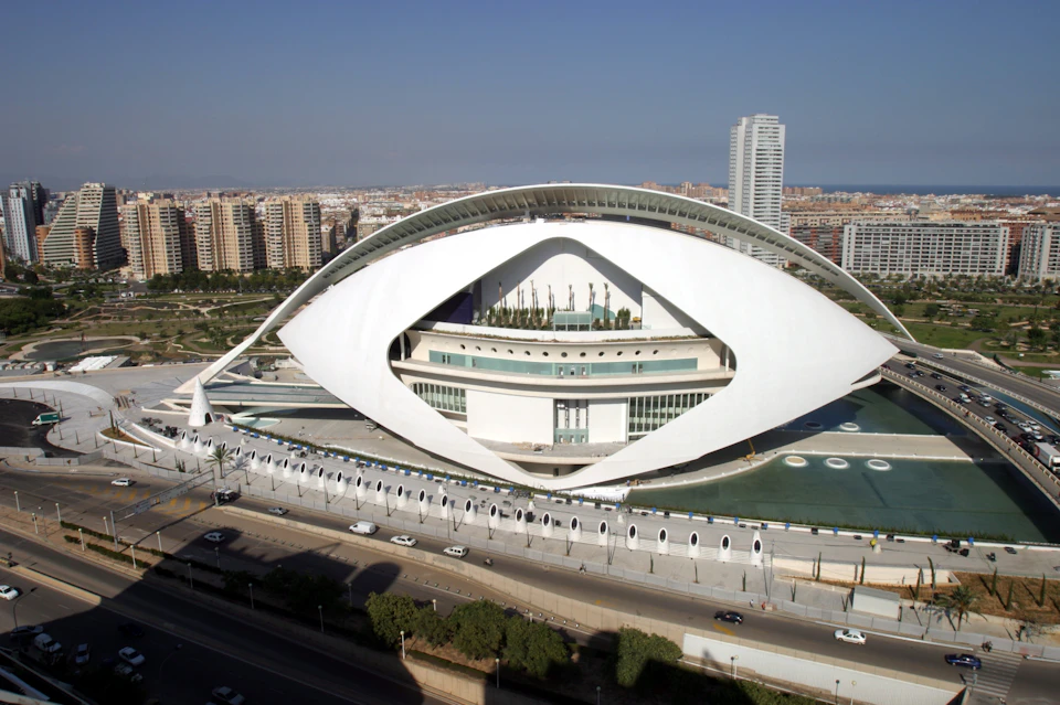 Futuristic white building with an eye-like shape, surrounded by water, with a cityscape and tall buildings in the background.