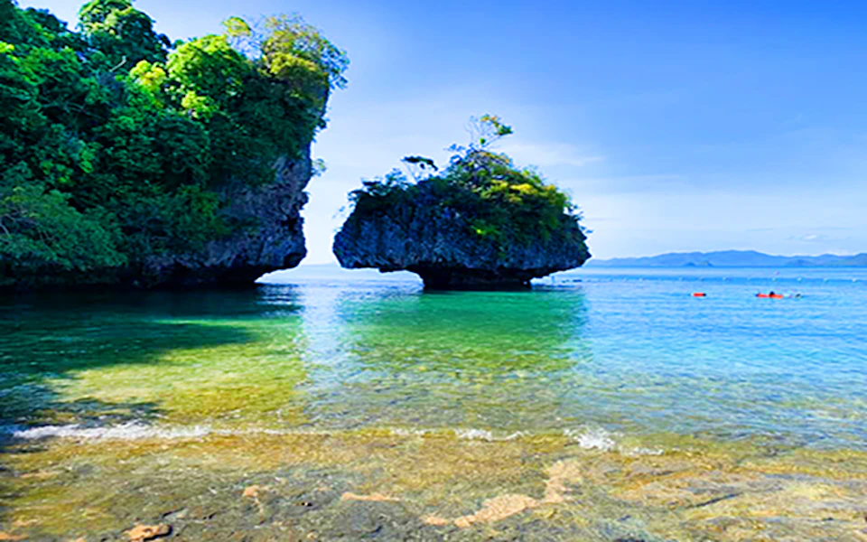 Clear blue water with two small, tree-topped rock formations in the distance under a bright, sunny sky.