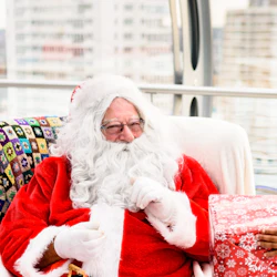 Santa Claus sits on a colorful blanket-covered chair, holding a present with a cityscape in the background.