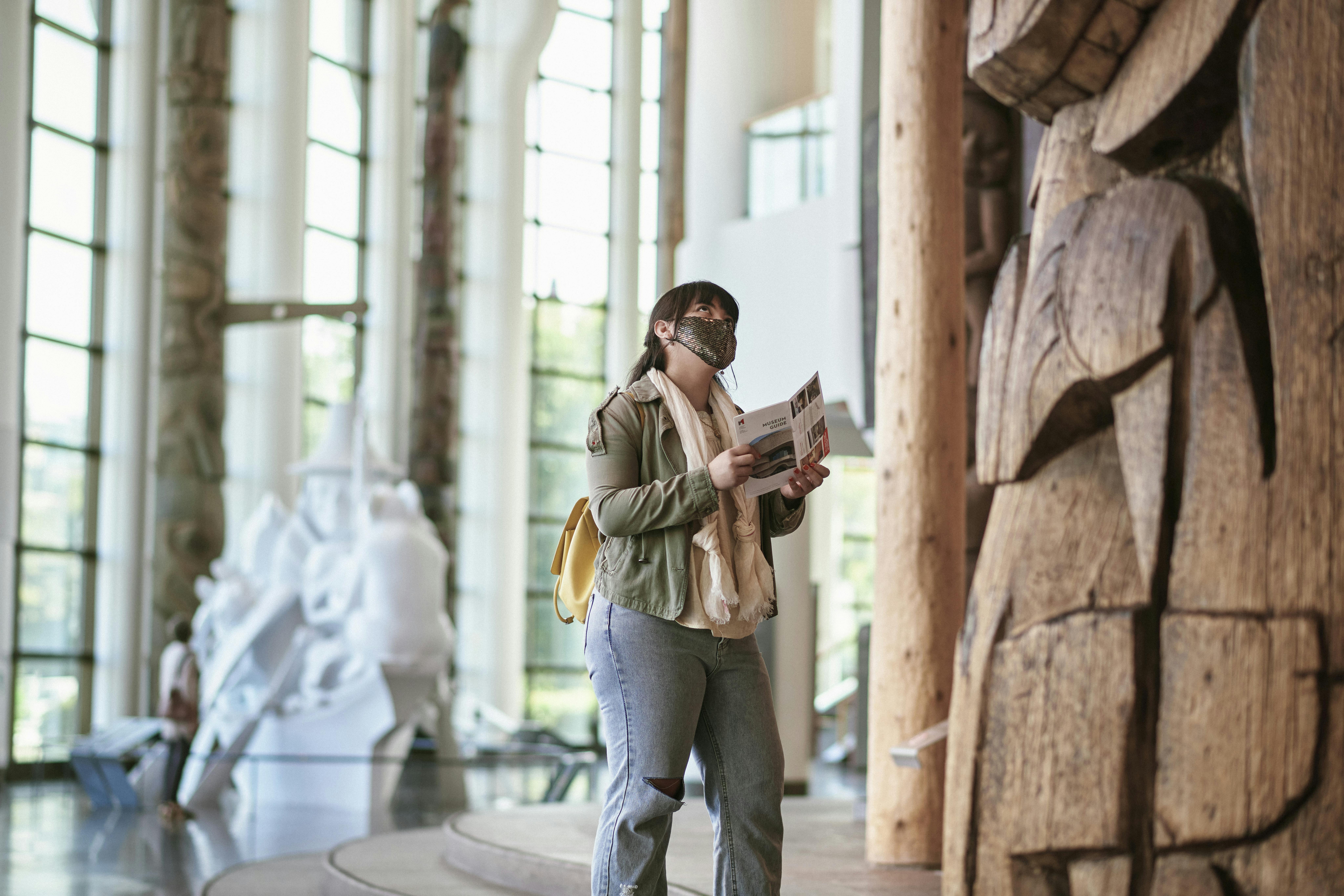 一个戴着口罩、拿着小册子的人站在一个宽敞、光线充足的博物馆里，附近有一座大型木制雕塑。