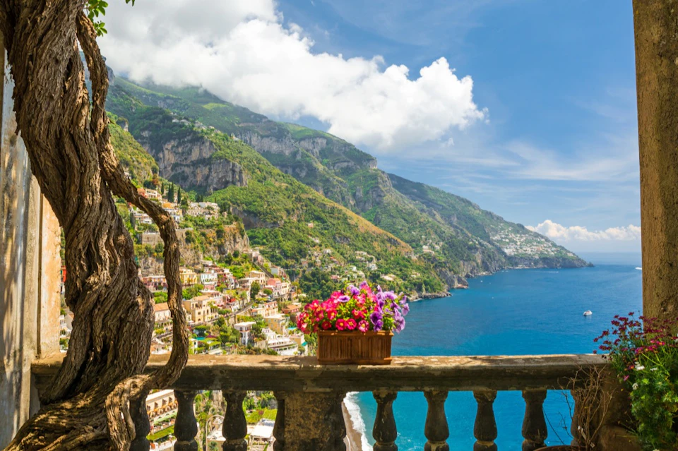 A scenic view of a coastal town with colorful buildings, lush green hills, and blue sea, seen over a balcony with flowers.