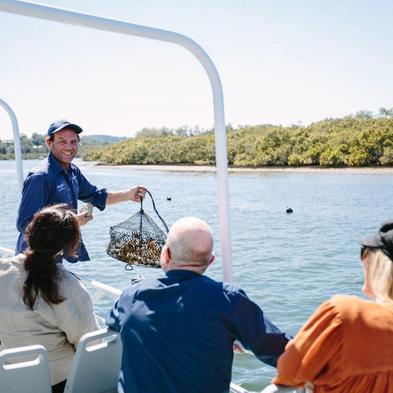 一位在船上微笑的男士向三个人展示了一张牡蛎网，坐在风景秀丽的河边，树木林立。